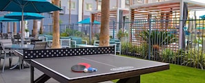 Une table de ping-pong installée à l’extérieur, avec un filet, entourée de chaises et de tables, avec des parasols et une clôture en arrière-plan.