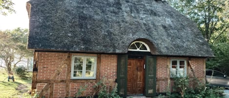 A small brick house with a thatched roof, a wooden door, and a stone pathway leading to the entrance.