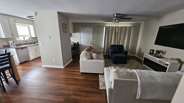 Una sala de estar con sofá, sillón y mueble para televisor. Hay un comedor con sillas y una mesa. El piso es de madera y tiene ventilador de techo.