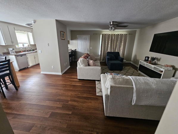 A living room with a sofa, armchair, and TV stand. There is a dining area with chairs and a table. The room has hardwood flooring and a ceiling fan.