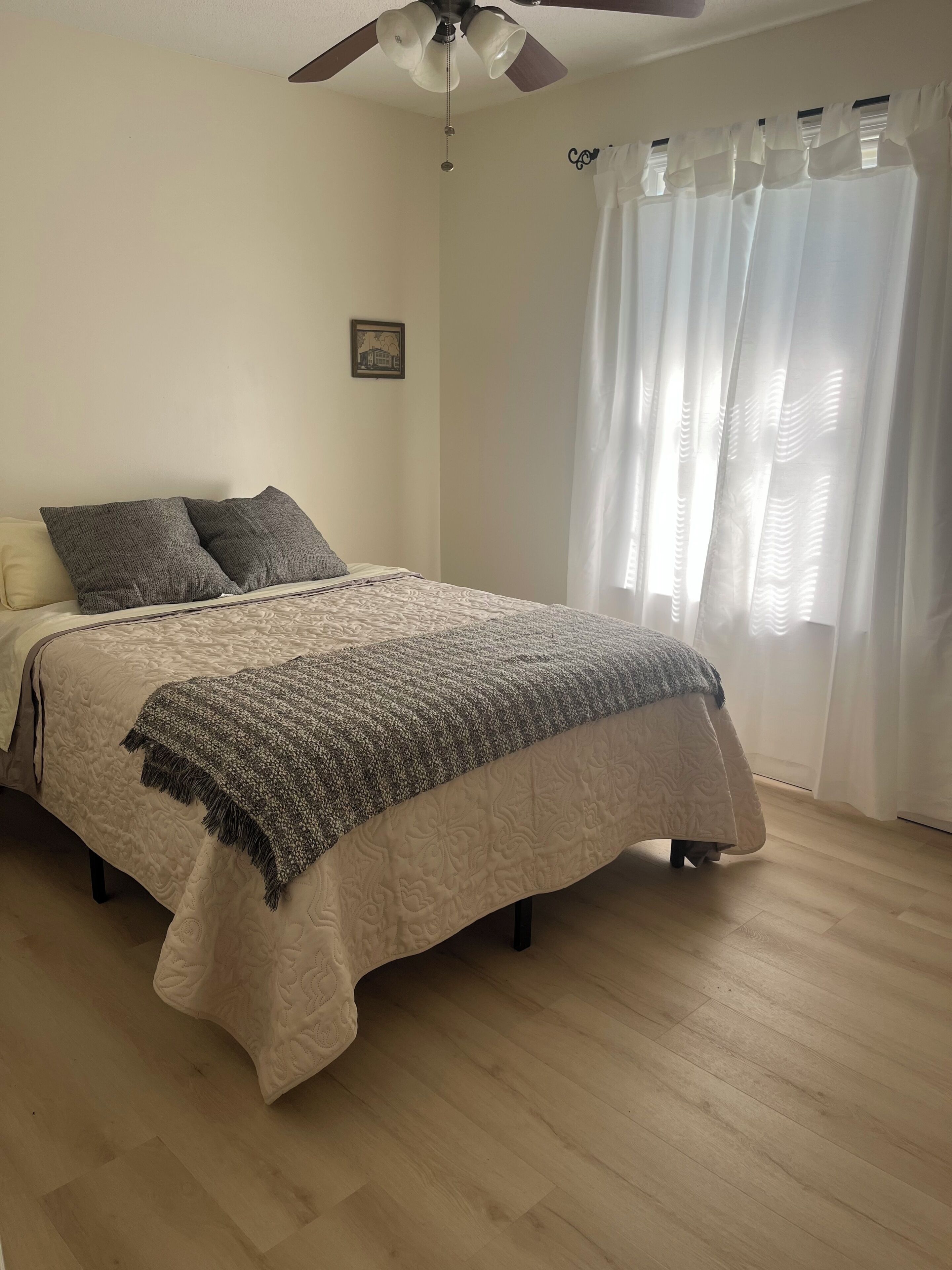 A bedroom with a bed, a ceiling fan, a window with curtains, and a framed picture on the wall.