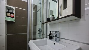 Un baño con un lavamanos blanco, cabina de ducha con mampara de vidrio y un estante con artículos de aseo.