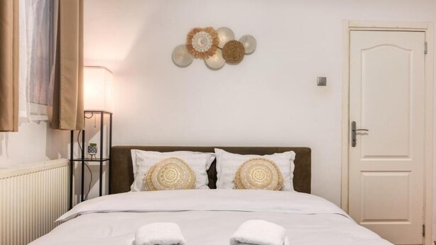 Una cama bien tendida con sábanas blancas y dos almohadas, una lámpara de noche y un adorno en la pared.