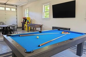 Ein Billardtisch mit Kugeln, ein Kicker und ein Fernseher in einem Raum mit Fenstern und Stühlen.