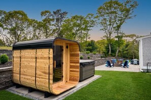 A wooden sauna with a curved roof, a built-in bench, and a patio area with outdoor furniture. - Lakewind House - sauna, hot tub, 5 min to Lake (Barnstable)
