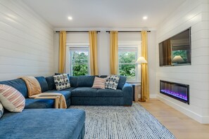 A modern living room with a sectional sofa, a fireplace, and large windows with curtains. - Lakewind House - sauna, hot tub, 5 min to Lake (Barnstable)
