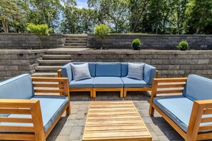 A patio with a sectional sofa, two wooden chairs, and a wooden table. - Lakewind House - sauna, hot tub, 5 min to Lake (Barnstable)