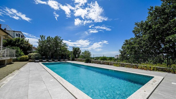 Une piscine rectangulaire, bordée d’une terrasse carrelée, entourée d’arbres et sous un ciel dégagé.