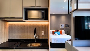 Quarto de hotel moderno com cozinha compacta, cama com almofadas e televisão.
