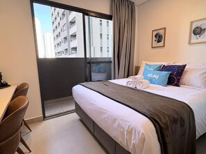 A hotel room with a bed, a desk, and a view of buildings through a window.