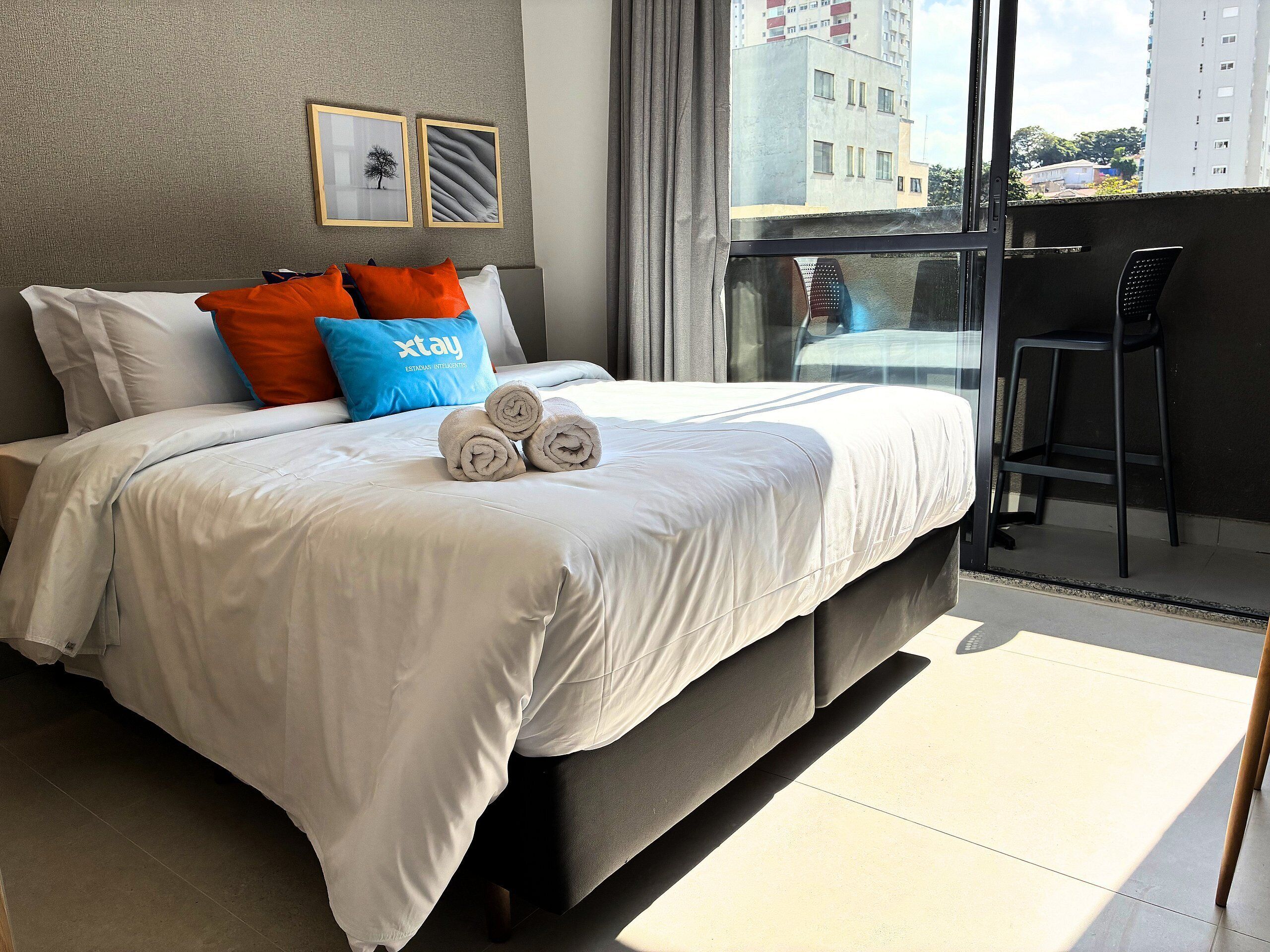 A modern hotel room with a large bed, a balcony with a view of the city, and a small table.