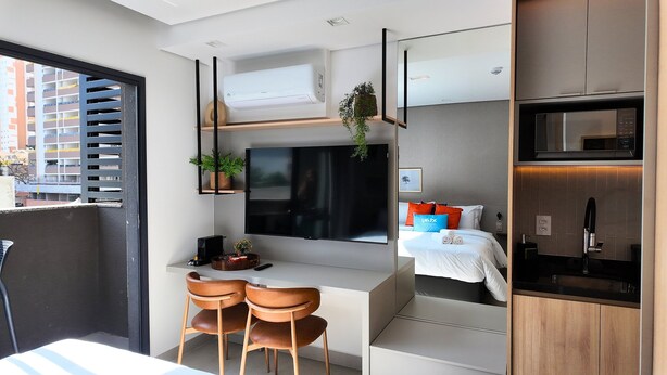 A compact hotel room with a kitchenette, a flat-screen TV, a bed with a nightstand, and a window overlooking a parking structure.