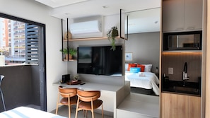 Quarto de hotel compacto com cozinha compacta, televisão de tela plana, cama com mesa de cabeceira e janela com vista para um estacionamento.
