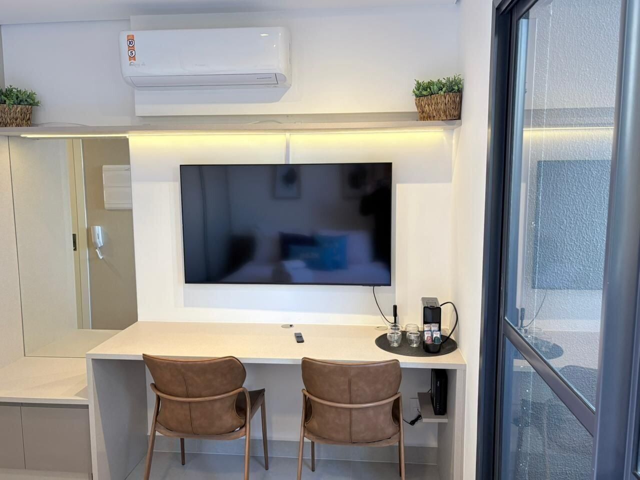 A modern room with a flat-screen TV mounted on the wall, a desk with two chairs, an air conditioning unit, and a window with a view.