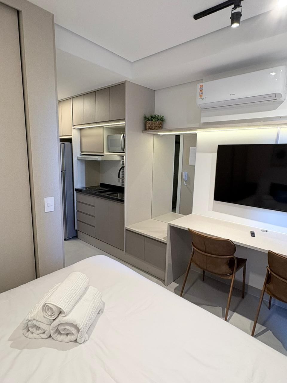 Een compacte hotelkamer met een bed, een bureau met stoel, een televisie, een kitchenette en een airconditioningsysteem.