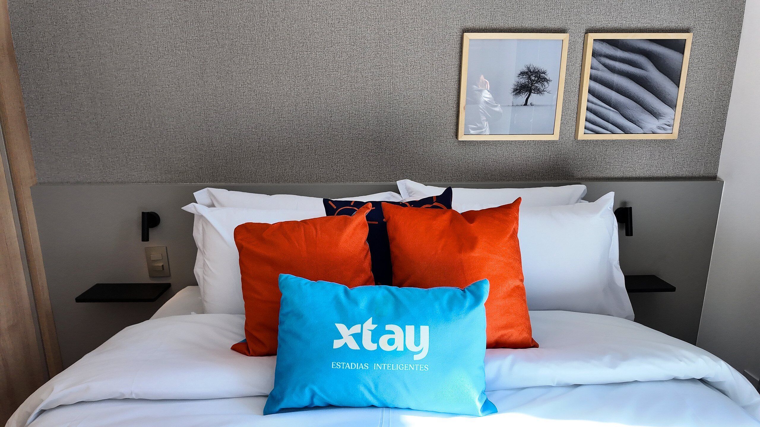 Una cama con ropa blanca, almohadas anaranjadas y azules, y dos cuadros en la pared.