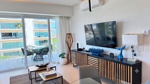 A modern living room with a large flat-screen TV, a glass coffee table, a sofa, a dining table, and a balcony with outdoor furniture. - Luxury Vallarta Marina 1BR, 180° Balcony & Golf Views (Puerto Vallarta)