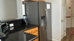 A kitchen with a stainless steel refrigerator, a black countertop, wooden cabinets, and a door with a white frame. - Beachside Vacation Home 117 #B (South Padre Island)