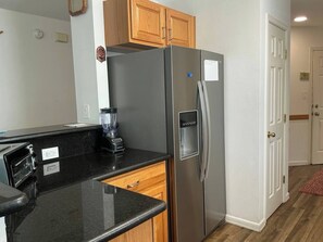 A kitchen with a stainless steel refrigerator, a black countertop, wooden cabinets, and a door with a white frame. - Beachside Vacation Home 117 #B (South Padre Island)