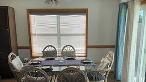 A dining area with a glass table, wicker chairs, and a window with blinds. - Beachside Vacation Home 117 #B (South Padre Island)