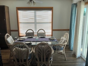 A dining area with a glass table, wicker chairs, and a window with blinds. - Beachside Vacation Home 117 #B (South Padre Island)