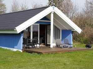 Et lille hus med en overdækket terrasse, en skydedør og en træterrasse med havemøbler.