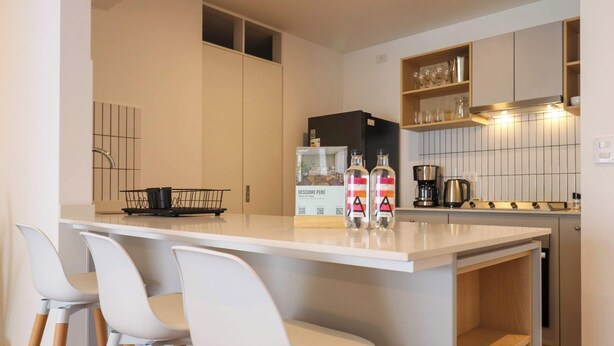 Cocina moderna con encimera blanca, taburetes de bar, cafetera y refrigerador.
