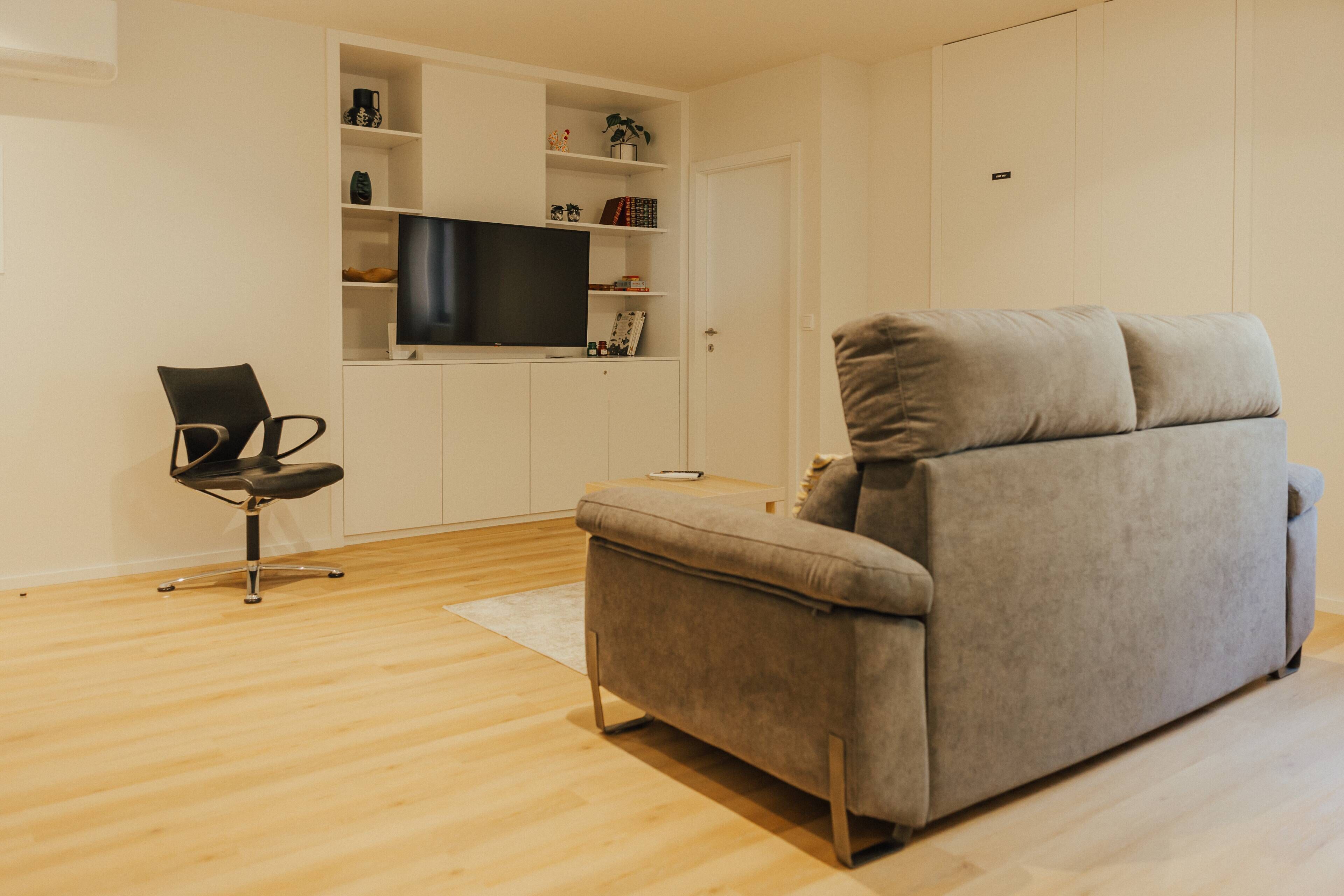 Un soggiorno moderno con un divano grigio, una poltrona da ufficio nera, una TV a schermo piatto montata su una parete bianca e un pavimento in legno.