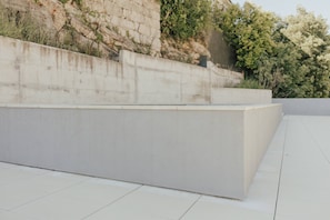 Une structure en béton dotée d’une surface plane, peut-être une terrasse ou une plateforme, située à côté d’un mur en pierre et d’une zone de végétation.