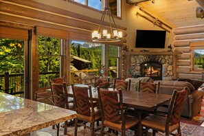 A log cabin interior with a stone fireplace, dining area, and large windows offering mountain views. - *New Listing* Big Sky Ski-In/Out Cabin w/ Hot Tub (Big Sky)