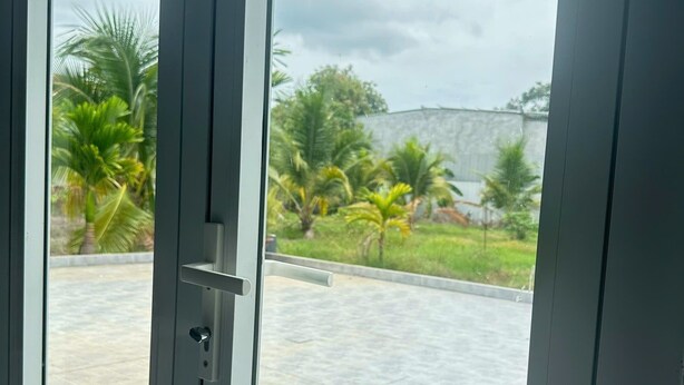 A view through a glass door with a metal handle, leading to a paved outdoor area with palm trees and a stone wall. - 3-bedroom house in welcoming Thanh Dien with AC comfort (Thanh Dien)