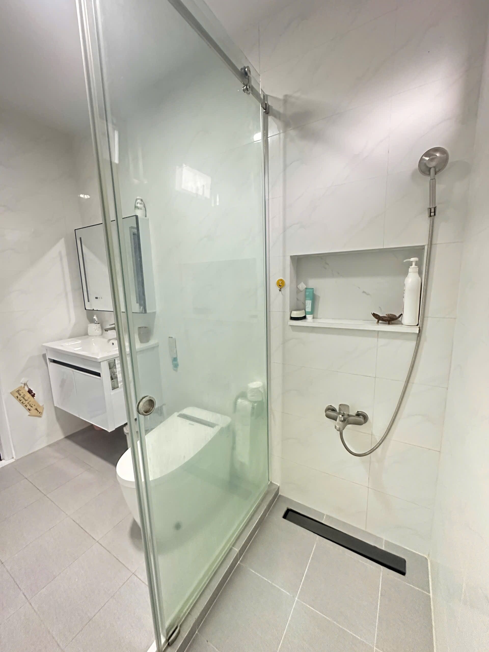 Un baño moderno con cabina de ducha de vidrio, inodoro, lavamanos y estante con artículos de aseo.