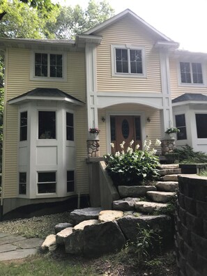 Ein zweistöckiges Haus mit einer Steintreppe zum Eingang, einem Erkerfenster links und einem Blumenkasten davor.