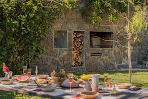 An outdoor dining area with a stone wall, a table set with food and drinks, and a view of a stone building.