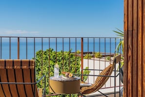 A balcony with a view of the sea, a chair, a small table with a bottle and food, and a wooden structure.