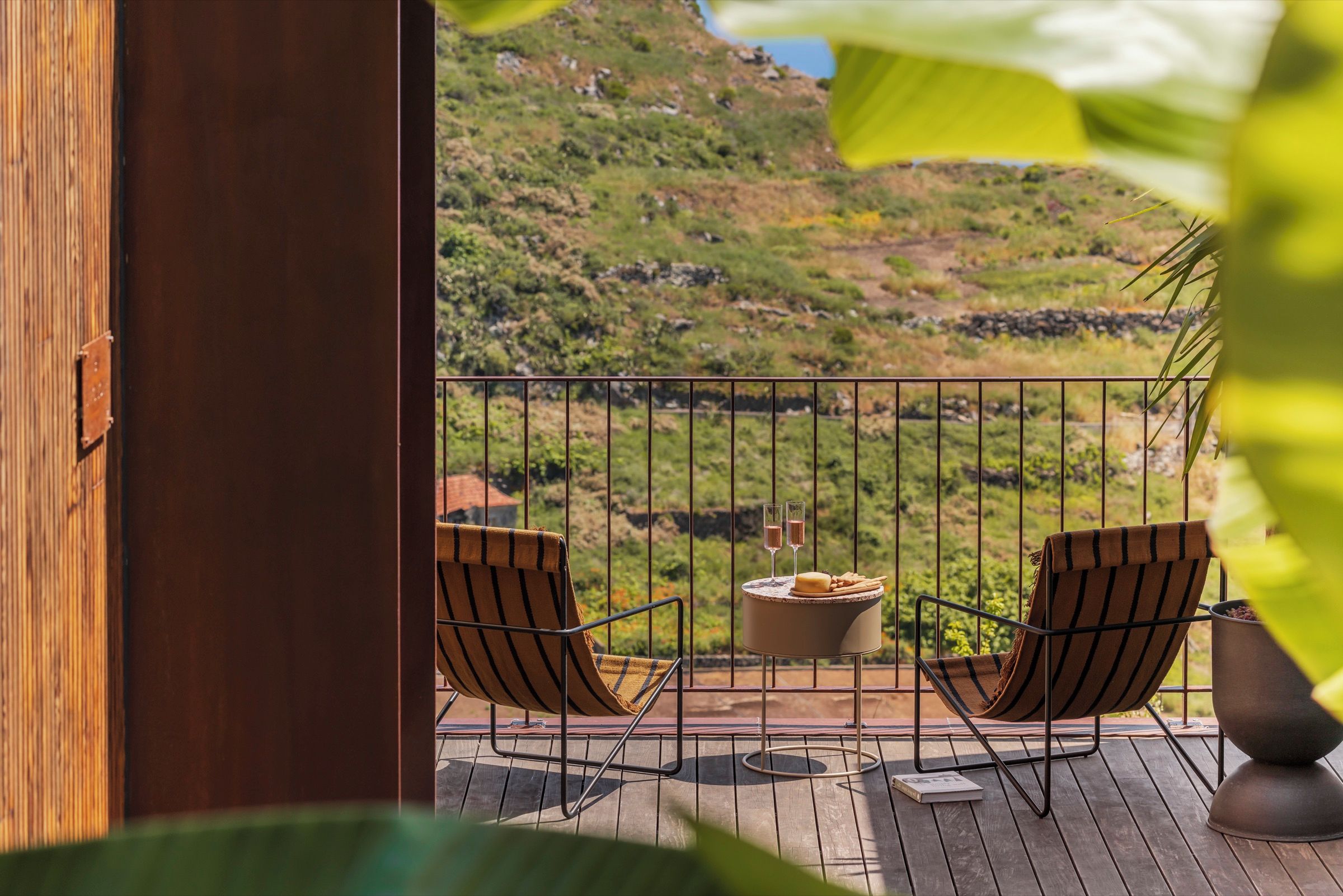 A balcony with two chairs, a table, and a view of a hilly landscape.