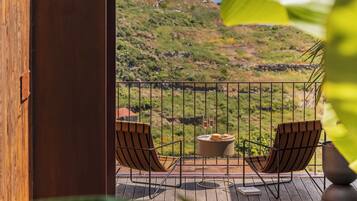A balcony with two chairs, a table, and a view of a hilly landscape.