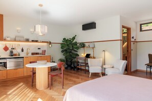 A modern hotel room with a bed, a dining table, chairs, a kitchen area, and a large potted plant.