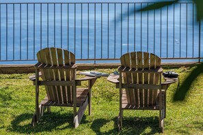 Dos sillas Adirondack con una mesa, un par de gafas de sol y una cámara sobre la mesa, situadas en un área verde con una baranda de metal y una masa de agua al fondo.