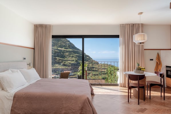 A hotel room with a bed, a dining table with chairs, a view of a mountainous landscape, and a balcony with a railing.