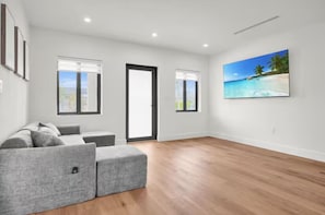 Ein geräumiges Wohnzimmer mit einer grauen Eckcouch, einem Flachbildfernseher an der Wand und Holzboden.