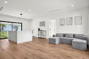 Ein modernes Wohnzimmer mit einem grauen Ecksofa, einer weißen Küchenzeile und einer Glastür, die auf einen Balkon führt.