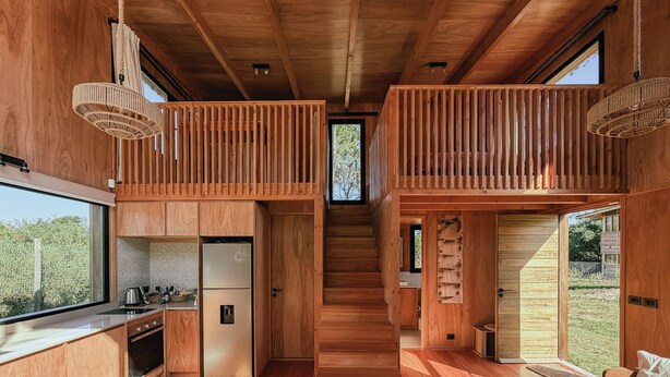 Um interior moderno em madeira, com cozinha, área de refeições e escada que leva a um mezanino.