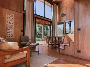 Ein geräumiges Zimmer mit Holzboden, einem Esstisch mit Stühlen, einer Couch und einem großen Fenster mit Blick ins Freie.