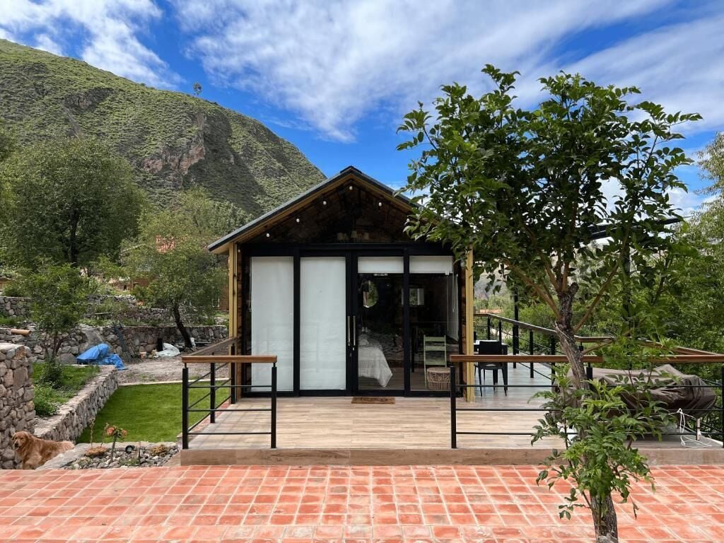 A small, modern building with a wooden exterior and large glass doors, surrounded by a garden and a mountain backdrop.