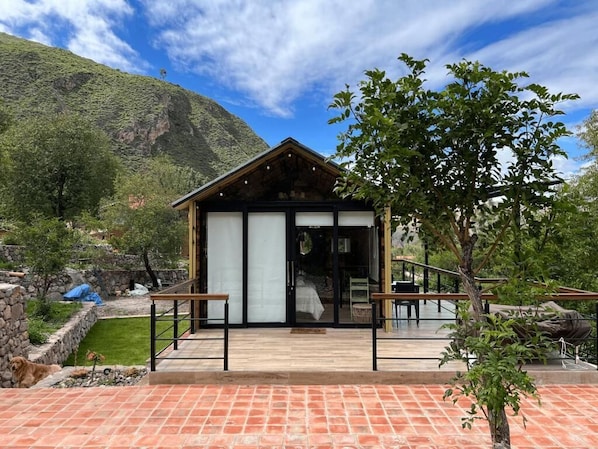 Un pequeño edificio moderno con fachada de madera y grandes puertas de cristal, rodeado de un jardín y con una montaña al fondo.