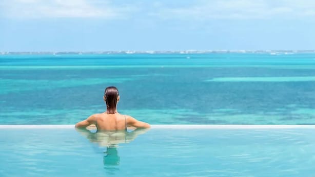 A person relaxing in a clear infinity pool with a view of the ocean and distant shoreline. - Luxury 2-bedroom Ocean view condo , Gym Spa & Infinity Pool (Cancún)