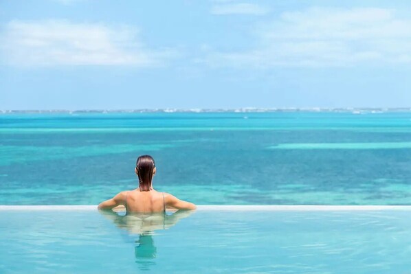 A person relaxing in a clear infinity pool with a view of the ocean and distant shoreline. - Luxury 2-bedroom Ocean view condo , Gym Spa & Infinity Pool (Cancún)