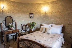 Un dormitorio con pared de piedra, cama con ropa de cama blanca, cómoda de madera, espejo y una silla.