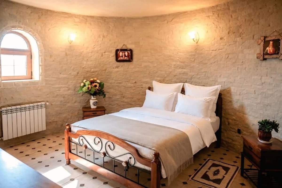 A bedroom with a stone wall, a bed with white linens, a nightstand with a vase of flowers, a small table with a plant, and a window with a view.